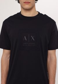 Camiseta de algodón negra con un logo cuadrado texturizado que presenta "A|X" y "ARMANI EXCHANGE BLUE EDITION" en gris. Mangas cortas, cuello redondo.