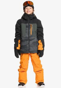 Veste de ski noire avec des accents gris et des zips orange, associée à un pantalon de neige orange ample et des bottes de ski noires.
