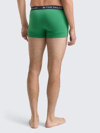 Uomo in piedi a piedi nudi, indossando boxer verdi Tom Tailor, visto di spalle su uno sfondo grigio chiaro uniforme.