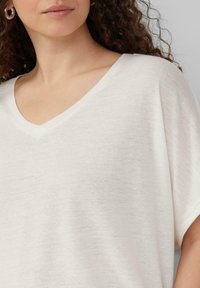 Luźna, biała bluzka z dekoltem w kształcie litery V, wykonana z lekkiego, teksturowanego materiału o delikatnym połysku. Krótkie, szerokie rękawy. Minimalistyczny design.