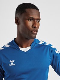 Blå sportsjersey med V-halsdesign, lavet af tekstureret stof. Har hvide chevron-detaljer på ærmerne og et lille logo på brystet.