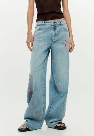 Széles szárú világoskék farmernadrág, színes hímzett csillagokkal és absztrakt mintákkal, fekete szandállal és ujjatlan barna felsővel viselve.