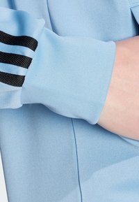 Světle modrý dlouhý rukáv oděv s texturovanou látkou. Obsahuje černé pruhované detaily na rukávech, detail manžety viditelný při bližším pohledu.