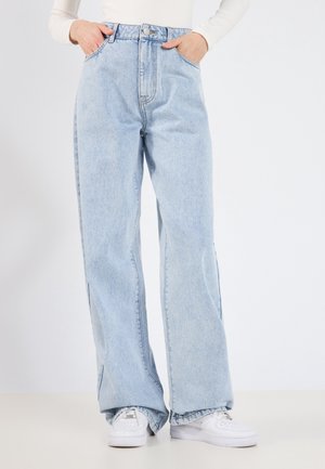 Person wearing light blue wide-leg jeans with hands in pockets and white sneakers against a plain white background.