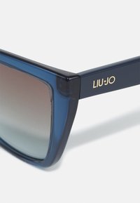 Solbriller med tykke, matte marineblå rammer, gradient brune linser og gull «LIU·JO» logo på siden, som fremhever et moderne geometrisk design.