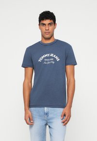 Vyras vilkintis tamsiai mėlyną Tommy Jeans marškinėlį su balta užrašu ir šviesiai mėlynomis džinsomis, stovi prieš paprastą baltą foną.