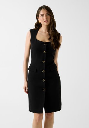 Kobieta z długimi, falującymi włosami, ubrana w czarną, bezrękawnikową sukienkę z złotymi guzikami i klapkowymi kieszeniami, stojąca na jednolitym jasnym tle.