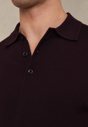 Detail osoby nosiacej tmavofialové polo tričko s dvoma zapnutými gombíkmi a zloženým golierom.