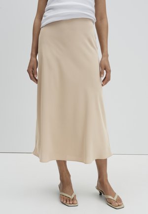 Frau mit einem beigen Satin-Midirock, weißem ärmellosem Oberteil und beigen Sandalen mit Absatz, stehend vor einem einfarbigen hellen Hintergrund.