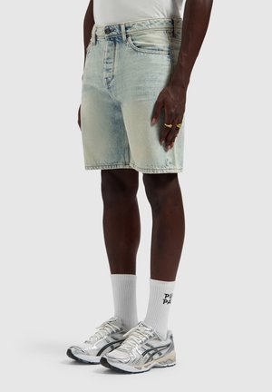 Lichtblauwe denimshorts gedragen met witte sneakersokken en zilveren hardloopschoenen door een persoon met een donkere huid, staand tegen een effen achtergrond.