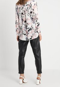 Blouse à manches longues blanche avec un motif floral en rose, noir et gris ; associée à un pantalon en cuir noir et des sandales blanches à talons blocs.