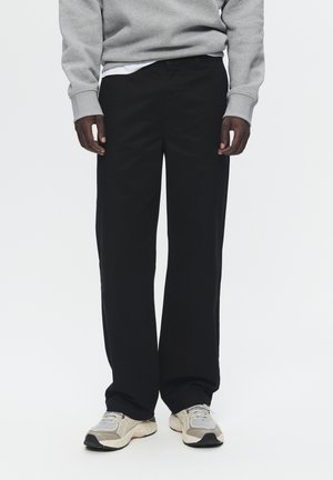 Black cotton trousers with a straight fit and slight sheen, paired with a grey sweatshirt and light beige sneakers, showcasing textured fabric.