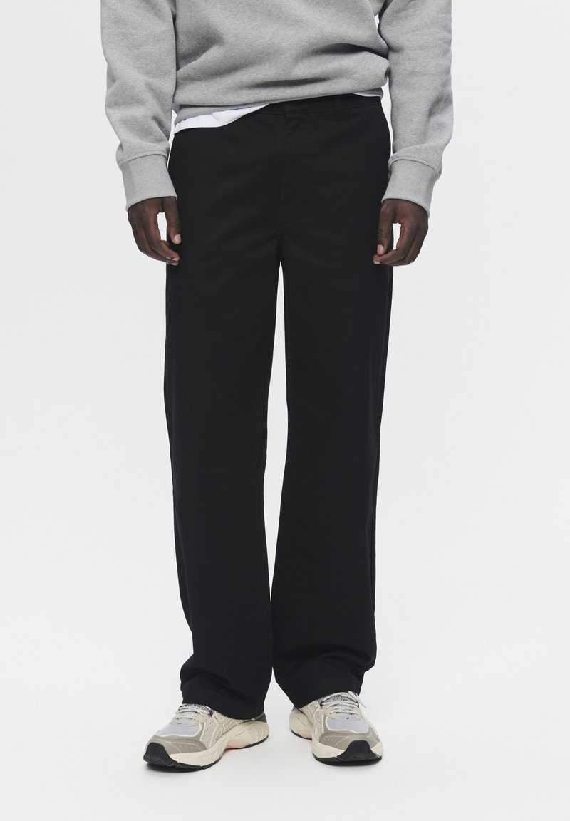 Black cotton trousers with a straight fit and slight sheen, paired with a grey sweatshirt and light beige sneakers, showcasing textured fabric.