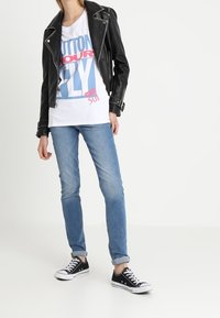 Chaqueta de cuero negro de motociclista, camiseta blanca con texto azul y rojo, jeans azul claro y zapatillas negras de caña baja con suela blanca, en pose de pie.