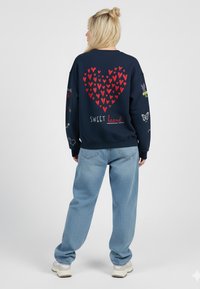 Felpa blu navy con motivo a cuore rosso e testo "SWEET heart" sul retro. Presenta vari dettagli grafici colorati sulle maniche.