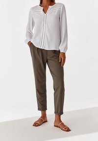 Blusa bianca a maniche lunghe con pois testurizzati e cuciture decorative, abbinata a pantaloni aderenti verde oliva e sandali bassi.
