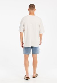 Světlé béžové oversized tričko s krátkými rukávy a modrým textem na levém rameni, kombinované s džínovými šortkami a hnědými sandály.
