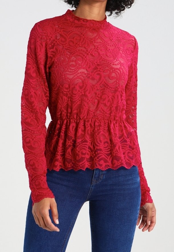 Femme portant un chemisier ajusté en dentelle rouge à manches longues avec un col haut et un ourlet à basque, associé à un jean bleu.