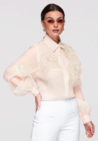 Femme portant un chemisier beige transparent avec des applications florales, un pantalon blanc, des boucles d’oreilles en forme de cœur et des lunettes de soleil teintées ambre, posant avec une main sur la hanche.