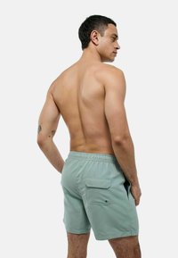 Shorts de bain vert clair avec une taille élastique, des poches latérales et une poche arrière avec rabat ; texture lisse, longueur mi-cuisse.