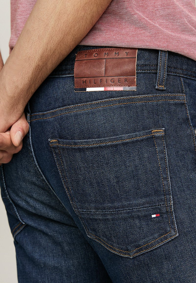 Dunkelblaue Denim-Jeans mit einem braunen Leder-Gürtel-Label, das "TOMMY HILFIGER" trägt. Sichtbare Nähte und ein kleiner farbiger Akzent an der Gesäßtasche.