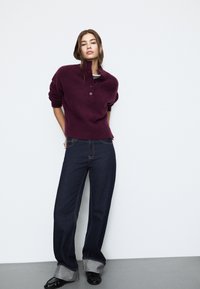 Bordo pleten pulover z visokim ovratnikom in tremi gumbi, v kombinaciji z temnimi širokimi kavbojkami z zavihanimi robovi in kontrastnimi šivi.