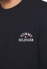 Camisa de algodón azul marino de manga larga con textura acanalada. Presenta el logo bordado en blanco "TOMMY HILFIGER" con acentos en rojo y blanco. Cuello redondo.