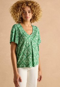 Zaļš īsām piedurknēm blūze ar V-veida kakla izgriezumu; tajā ir ziedu paisley raksts un brīvi plūstoša siluets. Stilizēta ar baltām pieguļošām biksēm.