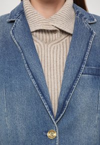 Denim sako u plavoj boji s izdubljenim reverima i jednim zlatnim kopčanjem, preko bež rebrastog pletenog duksera.