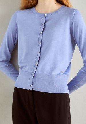 Donna che indossa un cardigan azzurro chiaro con bottoni e maniche lunghe e pantaloni neri, con le mani sui fianchi su uno sfondo semplice.