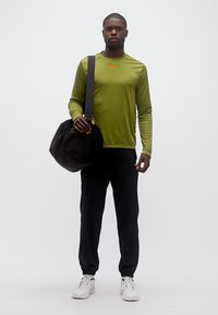 Νεαρός άνδρας που στέκεται, φορώντας μακρυμάνικο πουκάμισο ελιάς, μαύρο παντελόνι, λευκά αθλητικά παπούτσια, κρατώντας μια μαύρη τσάντα duffel στον ώμο του.