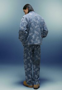 O persoană cu părul lung până la umeri, de culoare închisă, poartă o jachetă din denim albastru și pantaloni cu un model abstract alb, stând în fața unui fundal gradient albastru.