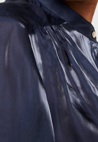 Blouse bleu marine à col montant, texture soyeuse et léger éclat, avec des détails plissés et une fermeture à bouton décoratif.