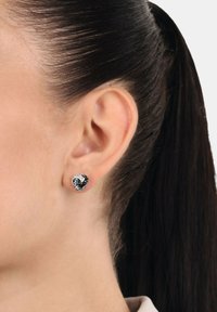 Kvinna med mörkt hår samlat bakåt, bärande en svart och silvrig vriden knopstudsörhänge i vänstra örsnibben mot en enkel bakgrund.