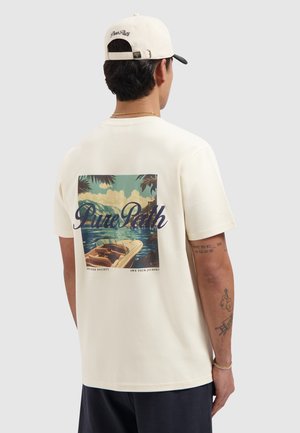 Man wearing cream t-shirt and cap, t-shirt back shows boat on lake with mountains, palm leaves, and text "Pure Path."
