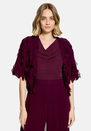 Vrouw met schouderlang blond haar draagt een donkerpaars getextureerd topje en bijpassende cape met gelaagde bladachtige stofdetails.