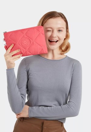 Quilted pink pouch with a circular logo, silver zipper, and fabric texture. Model holds it while wearing a gray long-sleeve shirt.