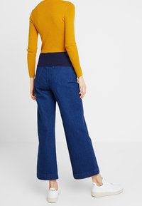 Yellow ribbed long-sleeve top with navy waistband, paired with wide-leg dark blue denim pants and white sneakers with clean lines.