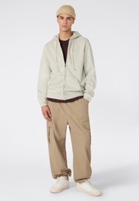 Sudadera con cremallera en gris claro y cordones, superpuesta a una camiseta oscura; combinada con pantalones cargo en color arena y zapatillas blancas. Paleta de colores neutros.
