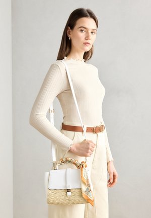 Jeune femme en haut côtelé beige et pantalon avec ceinture marron tenant un sac à main en paille et en cuir blanc avec une écharpe à motifs sur la poignée.