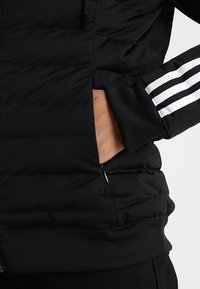 Svart vadderad jacka med en dragkedjeficka, ribbade muddarna och tre vita ränder längs ärmen. Slät, lätt tygtextur.