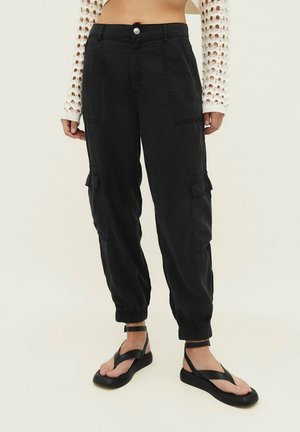 Person trägt schwarze Cargo-Hosen mit elastischen Bündchen, kombiniert mit schwarzen Riemchensandalen und einem weißen langärmeligen Häkelshirt.