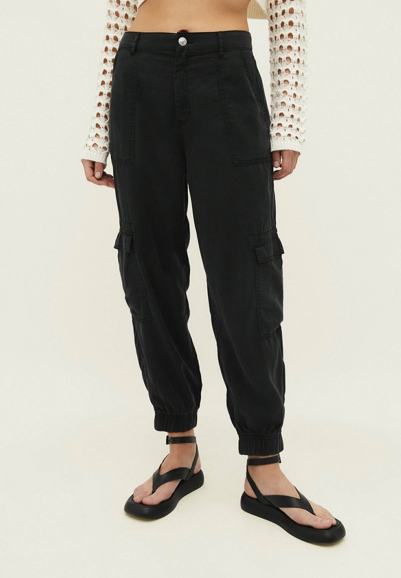 Personne portant un pantalon cargo noir avec des poignets élastiques, associé à des sandales noires à brides et un haut en crochet blanc à manches longues.