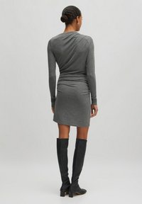 Graues langärmliges Jerseykleid mit figurbetontem Schnitt und gerafftem Rückendetail, kombiniert mit eng anliegenden schwarzen kniehohen Stiefeln.