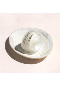 Barattolo di vetro bianco con testo nero e turchese su un piatto di ceramica bianco con bordi dorati. Crema liscia all'interno, posizionato su uno sfondo rosa pallido.
