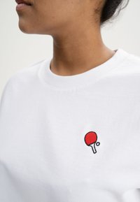 Witte katoenen t-shirt met een ronde hals, voorzien van een klein geborduurd rood tafeltennisbatje en bal op de linkerborst.