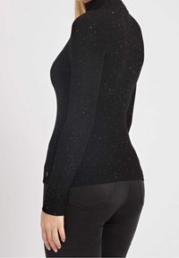 Maglione nero a collo alto a coste con spruzzi metallici, design aderente, maniche lunghe e dettagli del logo in modo sottile sull'orlo. Indossato con pantaloni scuri.