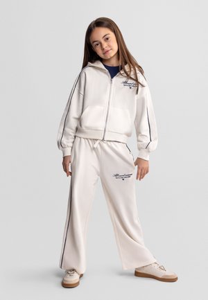 Młoda dziewczyna w białej bluzie z zamkiem i pasujących szerokich spodniach z granatowymi paskami po bokach oraz w trampkach, stojąca na szarym tle.