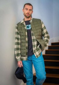 Gilet matelassé vert sur une chemise à carreaux, t-shirt noir et jean bleu. Une casquette sombre à la main, avec un fond neutre et des escaliers en bois.