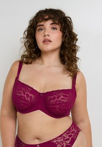 Donna dai capelli ricci che indossa un reggiseno di pizzo viola e mutandine di pizzo coordinate, in piedi davanti a uno sfondo grigio uniforme.
